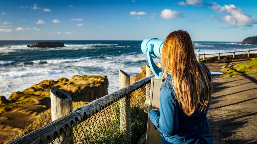 young woman looks through tourist telescope monocle at ocean waves, one of the best Oregon coast attractions