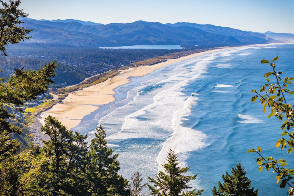An expansive, high-angle aerial view capturing the wide, sandy beach along the Tillamook Head trail, with the Pacific Ocean and surrounding coastal landscape under a clear, bright, sunny sky in Oregon