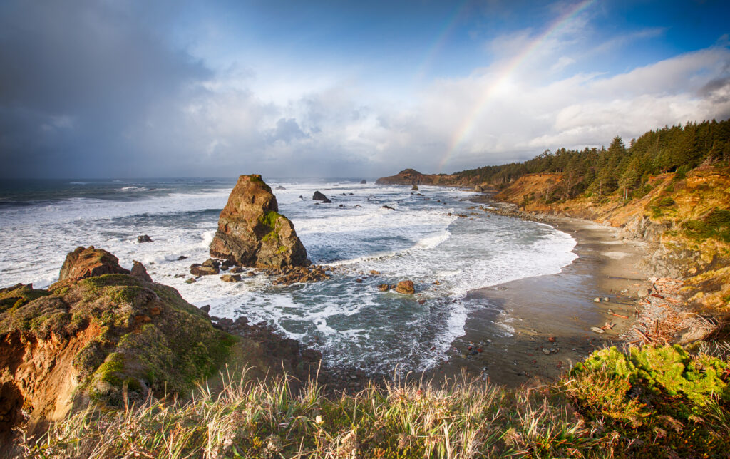 Rainbow in the background, dramatic sky, waves crashing onto shore at Gold Beach Oregon.