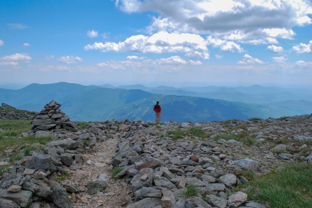 A man hiking the Presidential Range in New Hampshire, with a rocky trail and blue mountains in the backdrop.