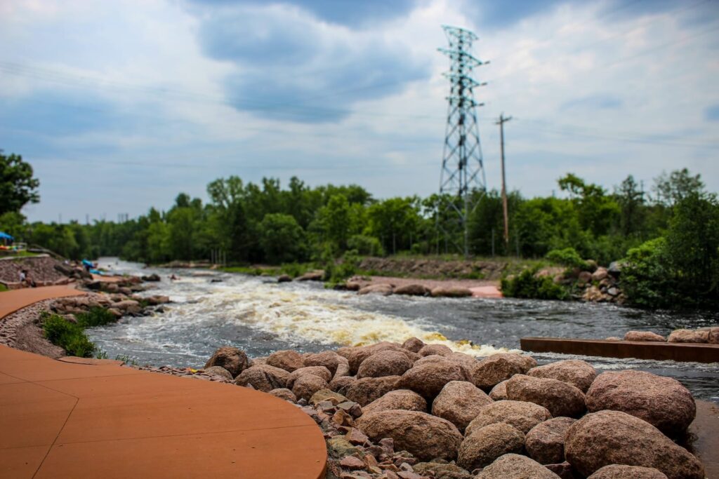 A river by a pathway, surrounded by trees and rocks, what you will see at the Wausau Whitewater Park.