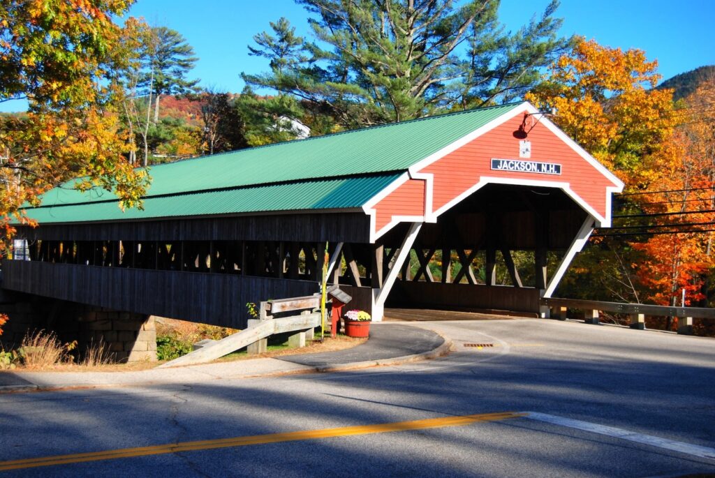 The Jackson NH Covered Bridge, red with a green roof, set among fall foliage, making it one of the best covered bridges in New Hampshire.