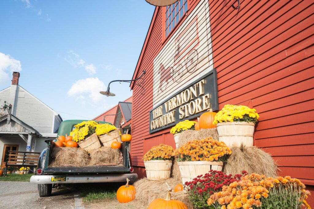 The Vermont Country Store sign on a red building surrounded by mums and pumpkins and an old truck, one of the best places to visit during Vermont in the fall.