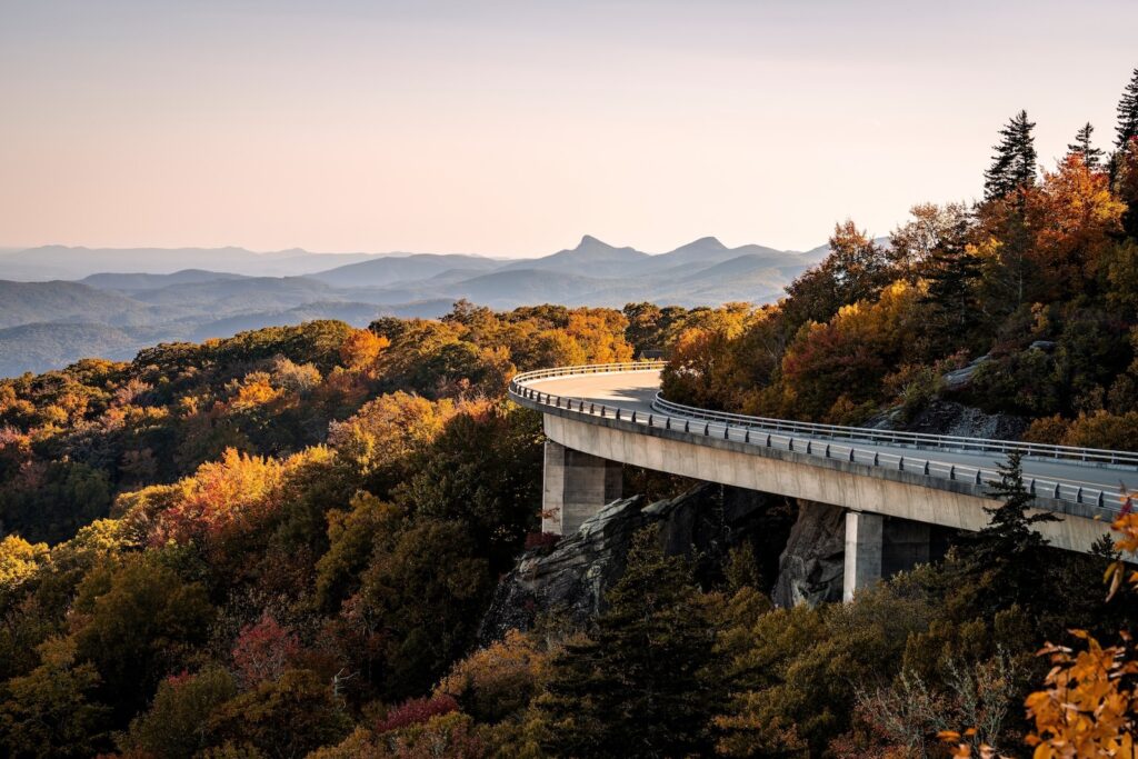 A viaduct bridge hugging a mountain of fall colors in North Carolina with blue mountains in the distance on the Blue Ridge Parkway.