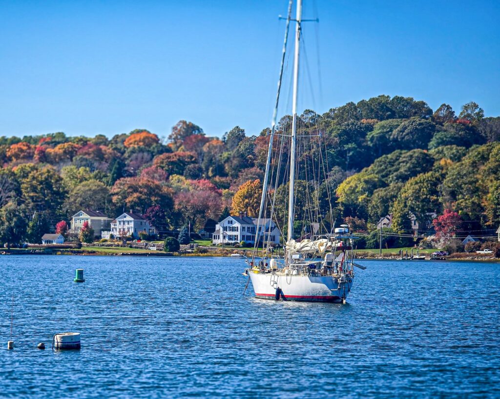 A sailboat on a river in front of fall foliage, what you can experience in Mystic on a weekend getaway in New England.