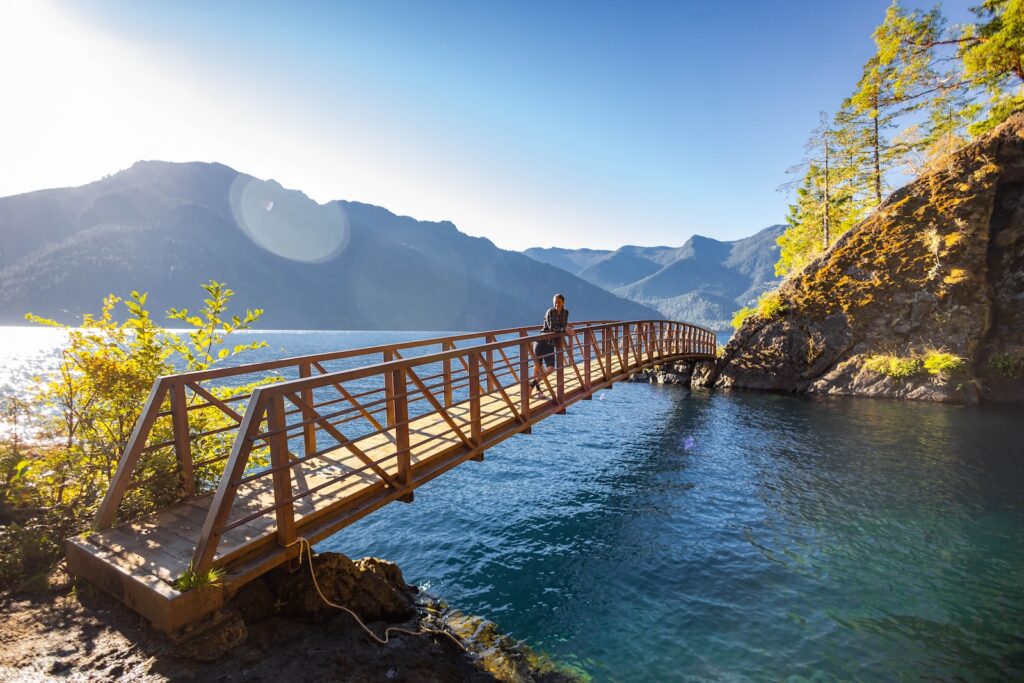 A girl standing on a bridge over water with mountains in the background at the Devil's Punchbowl at Lake Crescent, what you'll experience on the Spruce Railroad Trail in Olympic National Park.
