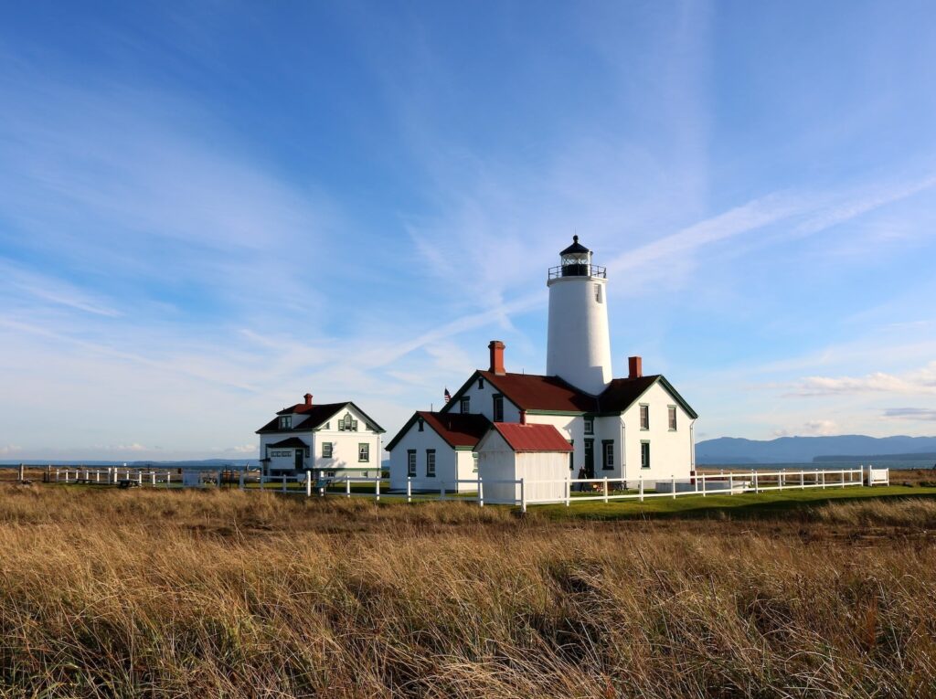 A white lighthouse with surrounding white buildings framed by a blue sky and grass, what visitors find when visiting the Dungeness Spit Lighthouse.