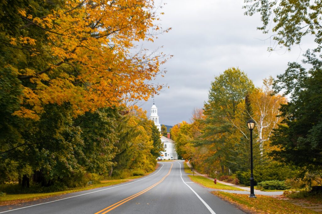 Route 100 Vermont running between fall foliage with a white steeple in the distance.