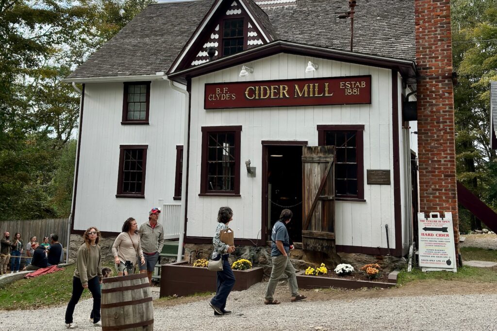 A white building with barrels, people, and mums out front at Clyde's Cider Mill in Mystic, CT.