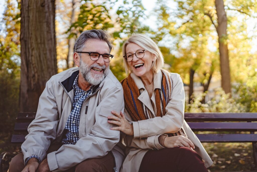 Happy senior couple sitting on a park bench in autumn, smiling and enjoying a sunny day outdoors with colorful leaves in the background. Here's how to Plan the Best Couples Getaway in Pennsylvania This Fall